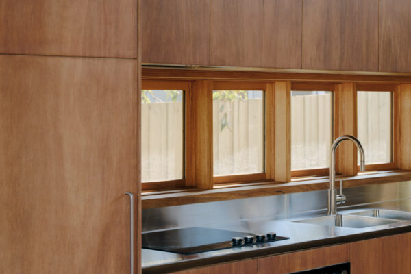 Kitchen in the Glaneuse project in Point Lonsdale, Victoria, featuring timber joinery, a stainless steel benchtop and splashback, timber-framed highlight windows, and integrated appliances.