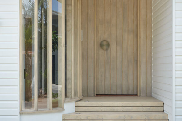 Entry porch in the Dylan Lane family home renovation in Byron Bay, featuring curved timber-framed glazing, vertical timber cladding, a recessed front door and a soft coastal material palette.