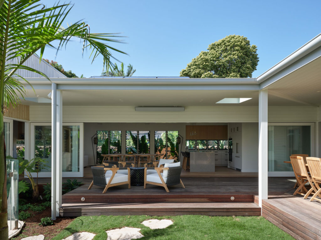 Rear outdoor living area in the Dylan Lane family home renovation in Byron Bay, featuring a covered timber deck, open-plan indoor-outdoor connection, white weatherboard cladding and landscaped garden surrounds.