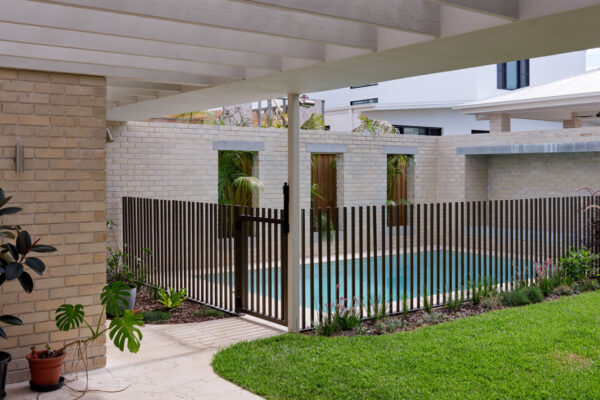 Pool and landscaped courtyard in the Sunhaven Avenue home build in Skennars Head, featuring light brick walls, vertical metal pool fencing, tropical planting and a covered outdoor area.