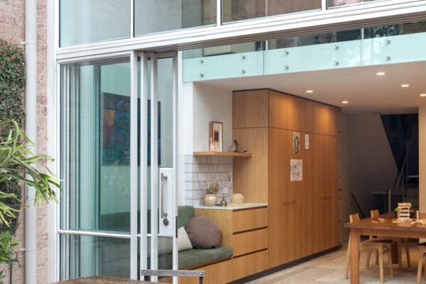 Open-plan kitchen and dining area in the Park House build in Parkville, Melbourne, featuring timber joinery, white cabinetry, double-height glazing and layered connected living spaces.
