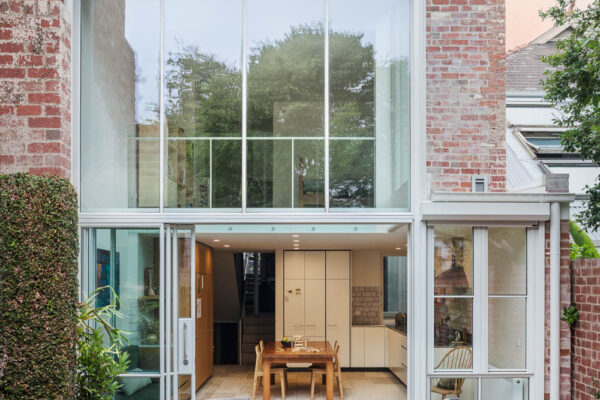 Open-plan kitchen and dining area in the Park House build in Parkville, Melbourne, featuring timber joinery, white cabinetry, double-height glazing and layered connected living spaces.